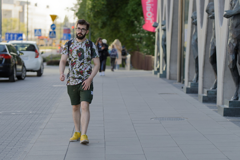 18 - A man walking down a paved sidewalk wearing a patterned t-shirt, dark shorts, and yellow shoes